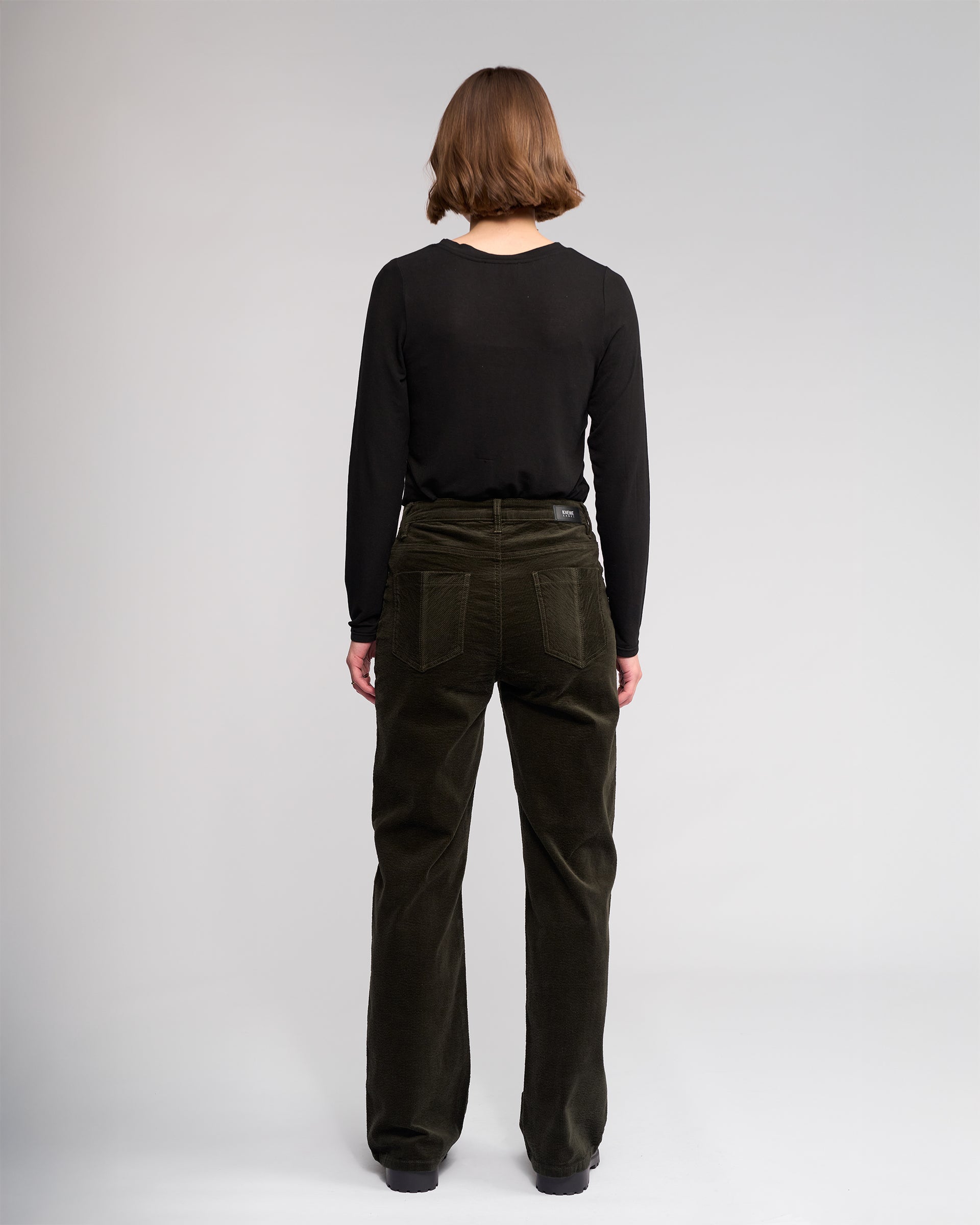 A person with short brown hair faces away, wearing a black long-sleeve shirt and Knewe Label’s Dallas Cord - Hunter wide-leg pants, against a plain light gray background.