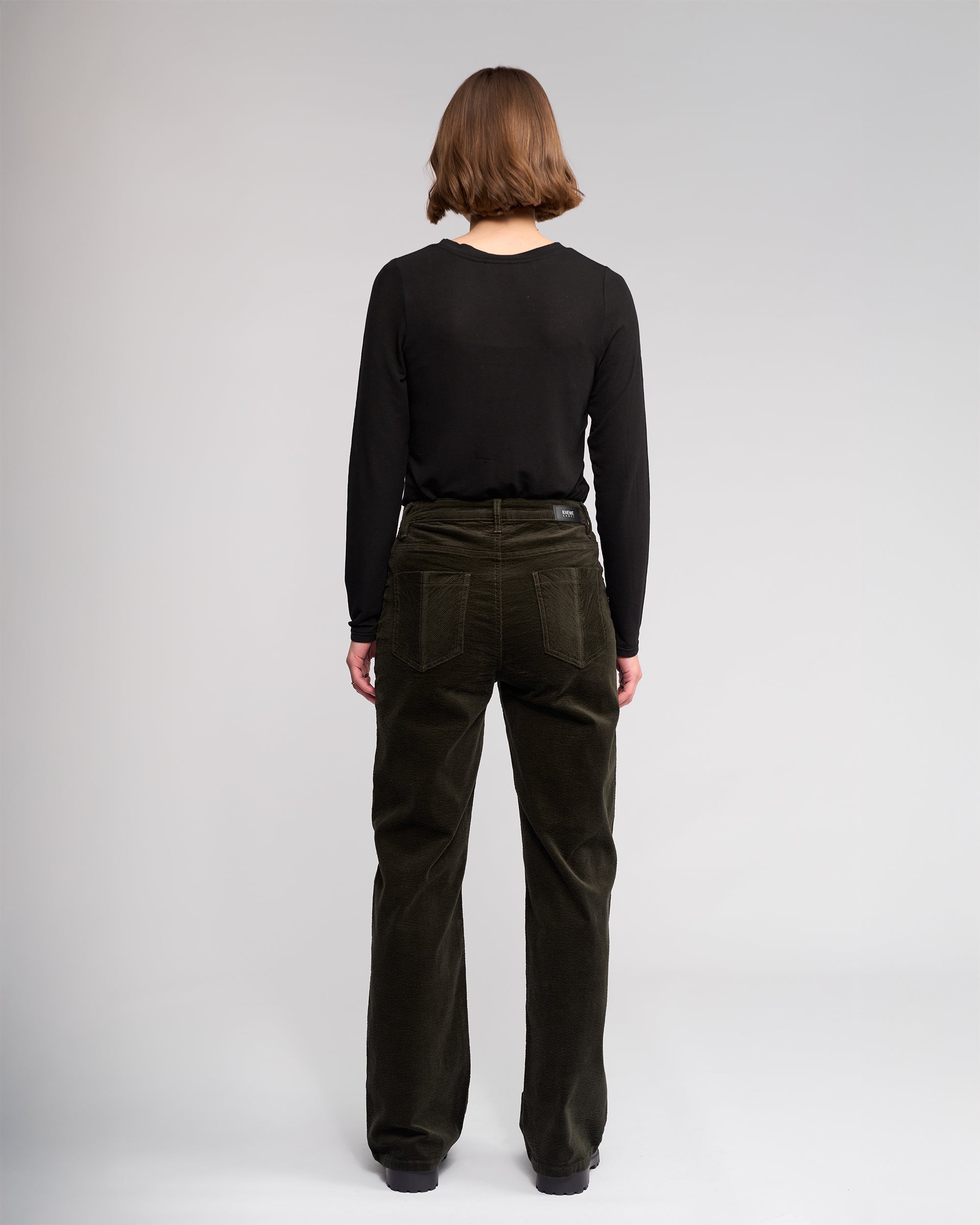 A person with short brown hair faces away, wearing a black long-sleeve shirt and Knewe Label’s Dallas Cord - Hunter wide-leg pants, against a plain light gray background.