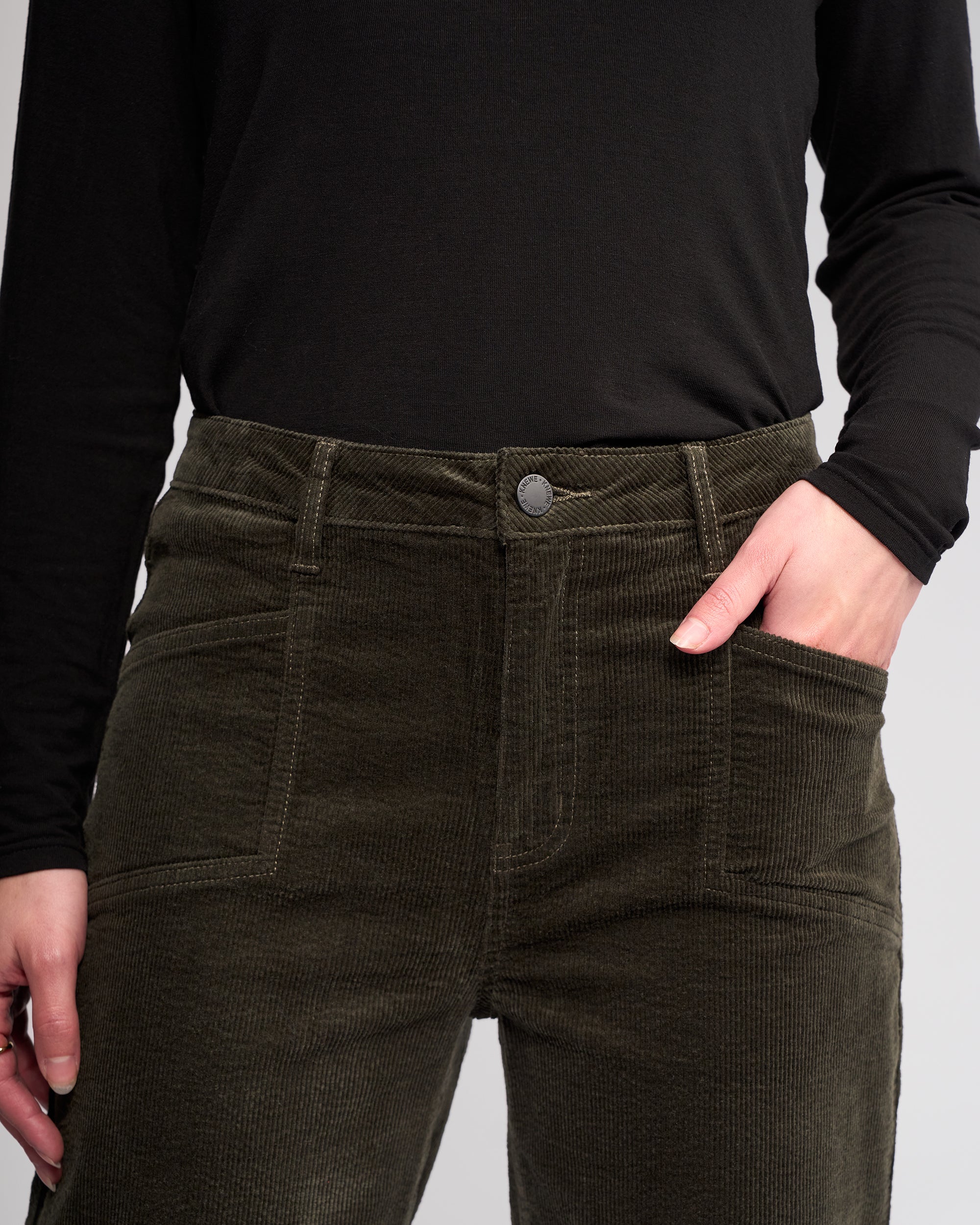A person wearing Knewe Label’s Dallas Cord - Hunter wide-leg pants and a black long-sleeve shirt stands with one hand in their pocket against a neutral background.