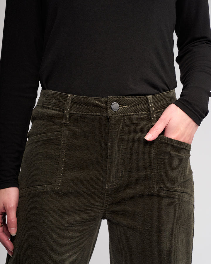 A person wearing Knewe Label’s Dallas Cord - Hunter wide-leg pants and a black long-sleeve shirt stands with one hand in their pocket against a neutral background.