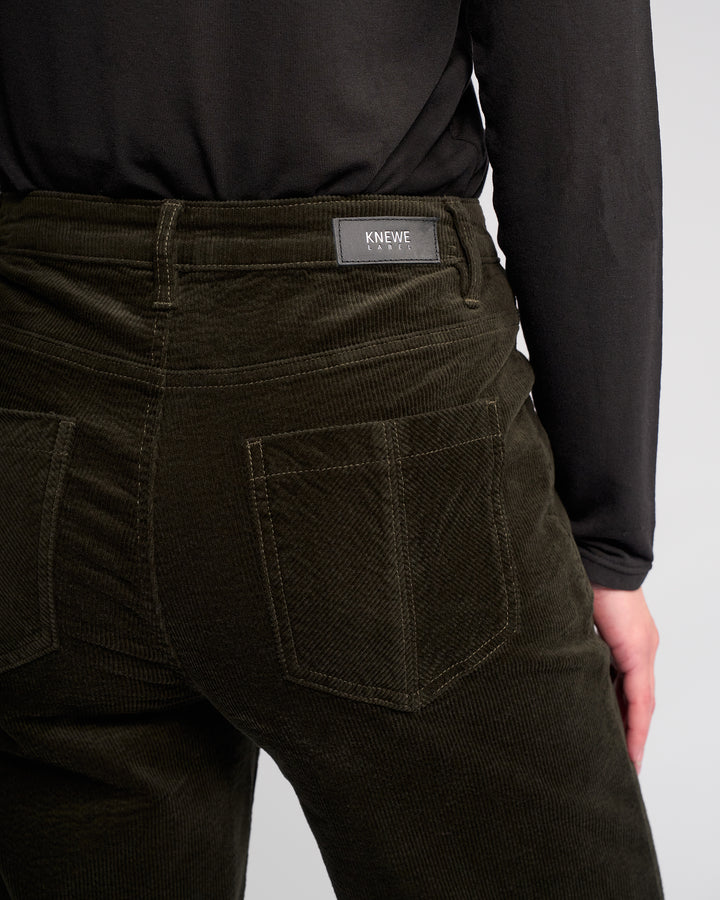A person shown from the back wears Dallas Cord - Hunter wide-leg corduroy pants by Knewe Label in dark green, paired with a black top. The full-length pants have large back pockets and a Knewe Label patch on the waistband.