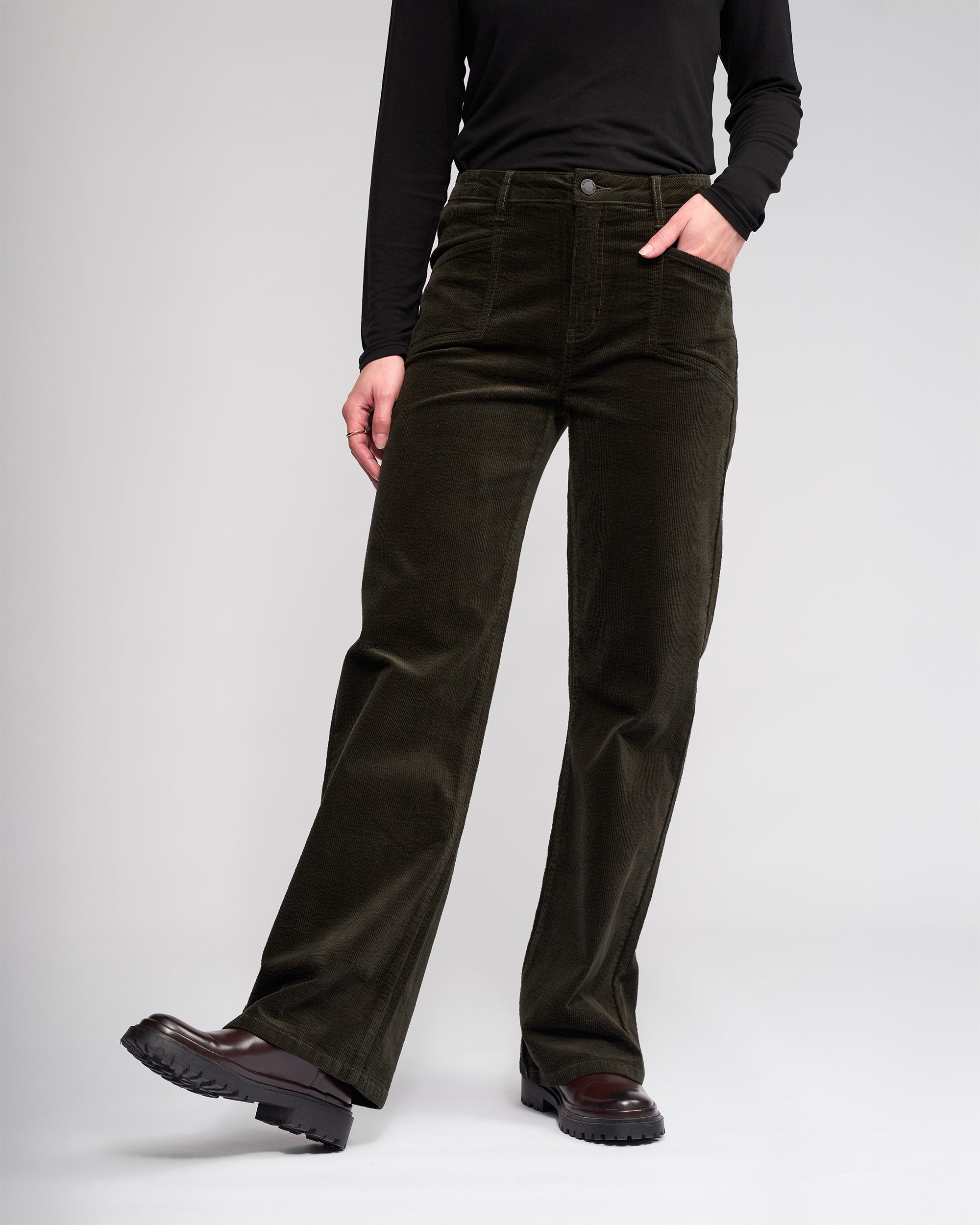 A person wearing a long-sleeve black top, Dallas Cord - Hunter wide-leg pants from Knewe Label, and black chunky shoes stands with one hand in their pocket against a plain light background.