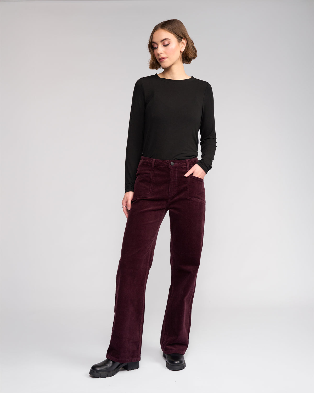Woman stands against a plain background wearing Knewe Label’s Dallas Cord - Raisin wide-leg pants, a black long-sleeve top, and black shoes, one hand in her pocket as she looks slightly to the side.