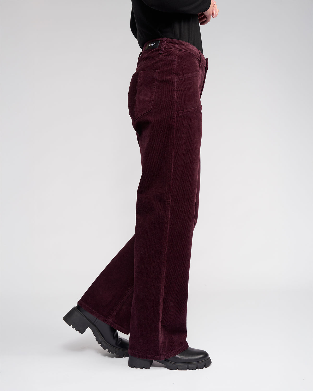 Someone stands sideways against a plain light background, wearing Knewe Label's Dallas Cord - Raisin, a high-waisted, wide-leg corduroy pant paired with black chunky-soled shoes.