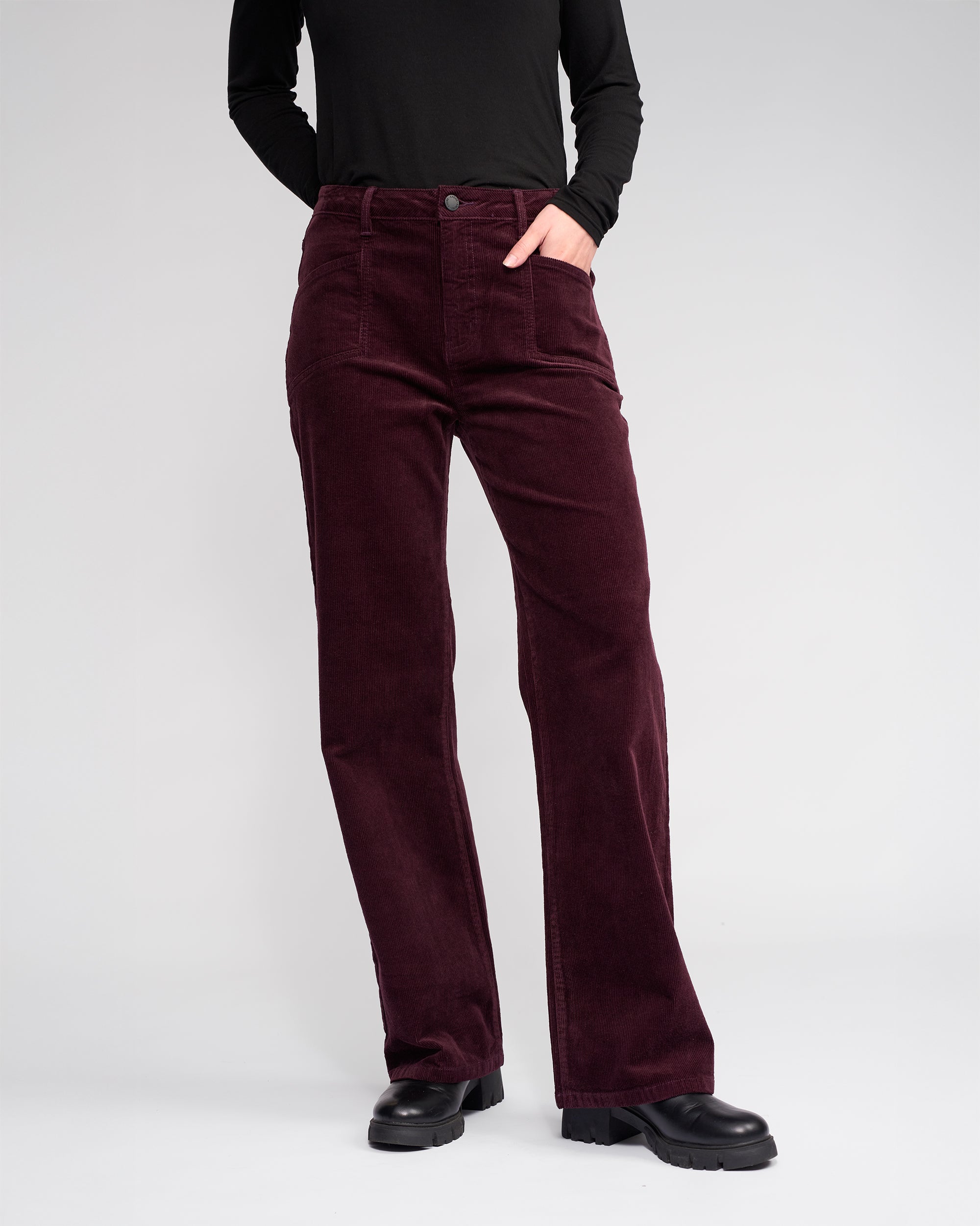 Wearing Knewe Label’s Dallas Cord pants in raisin and a black long-sleeve top with black shoes, a person stands against a light background, one hand in their pocket.