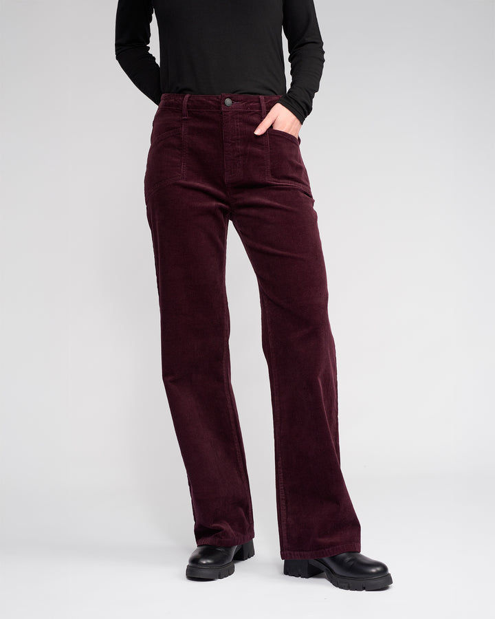 Wearing Knewe Label’s Dallas Cord pants in raisin and a black long-sleeve top with black shoes, a person stands against a light background, one hand in their pocket.