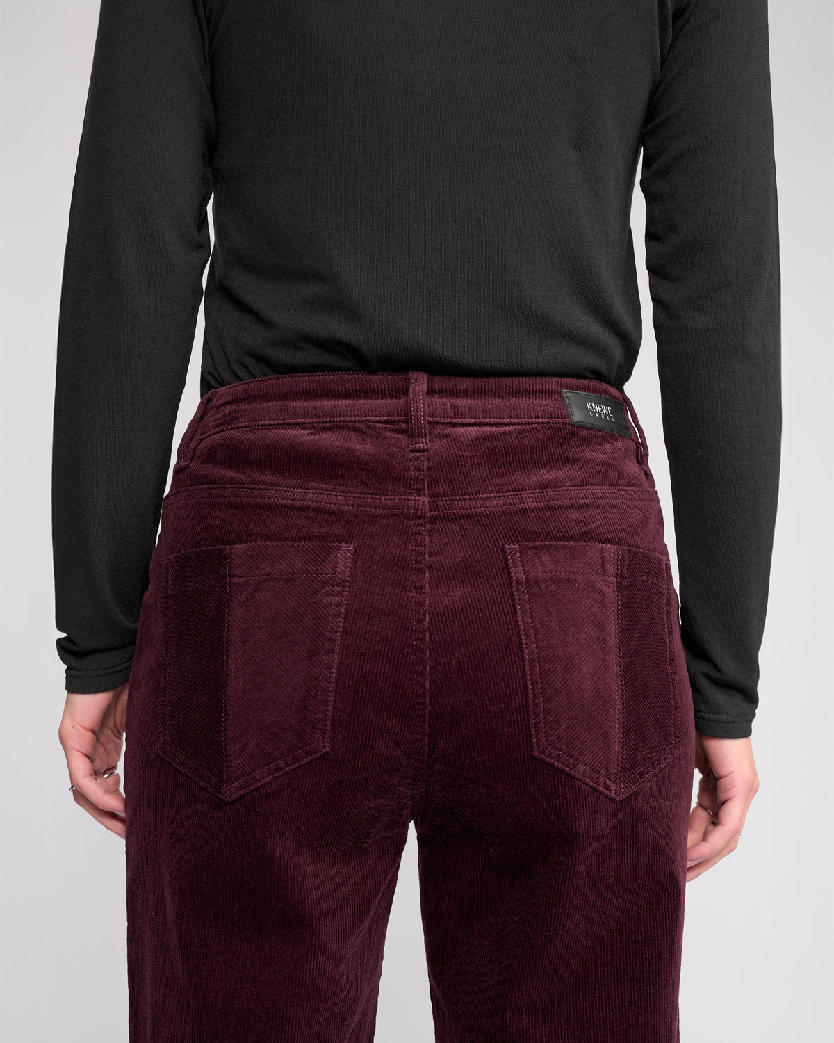 Person wearing the Knewe Label Dallas Cord wide-leg pant in Raisin, seen from the back. The pants feature two back pockets and are paired with a black long-sleeve top. Hands rest relaxed by their sides.