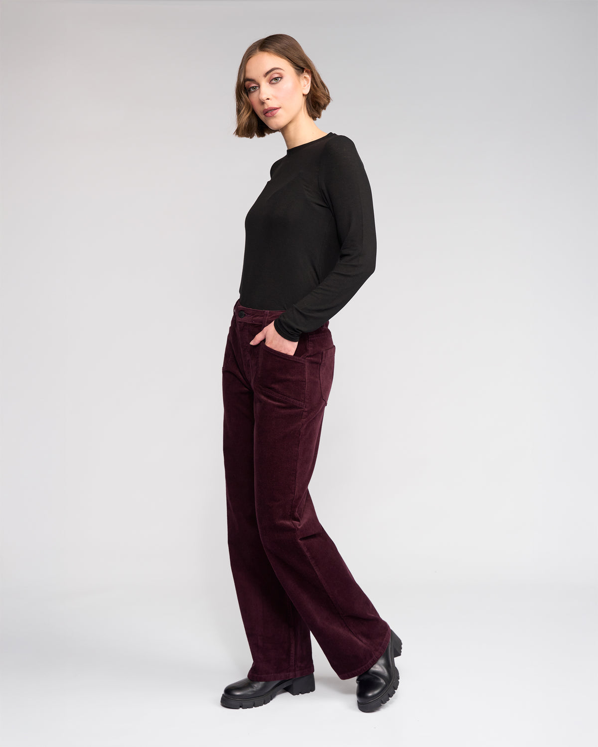 A woman with short brown hair stands with her hands in her pockets, wearing Knewe Label’s Dallas Cord - Raisin wide-leg corduroy pants and a black long-sleeve top, paired with black chunky shoes against a plain background.