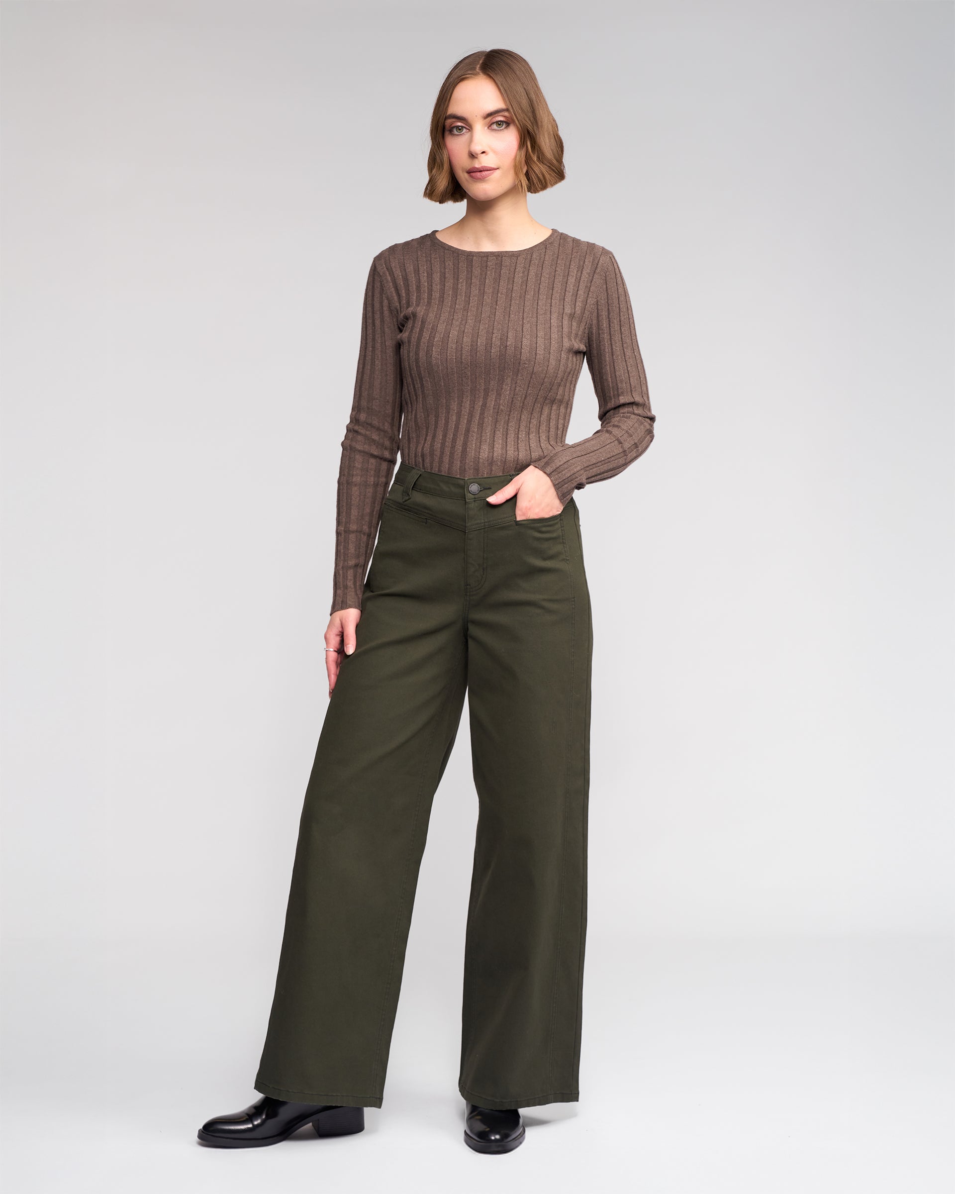 A woman with short brown hair and a neutral expression wears the Knewe Label Hollie Jean - Hunter, paired with a long-sleeved brown ribbed top and black shoes, standing before a plain background.