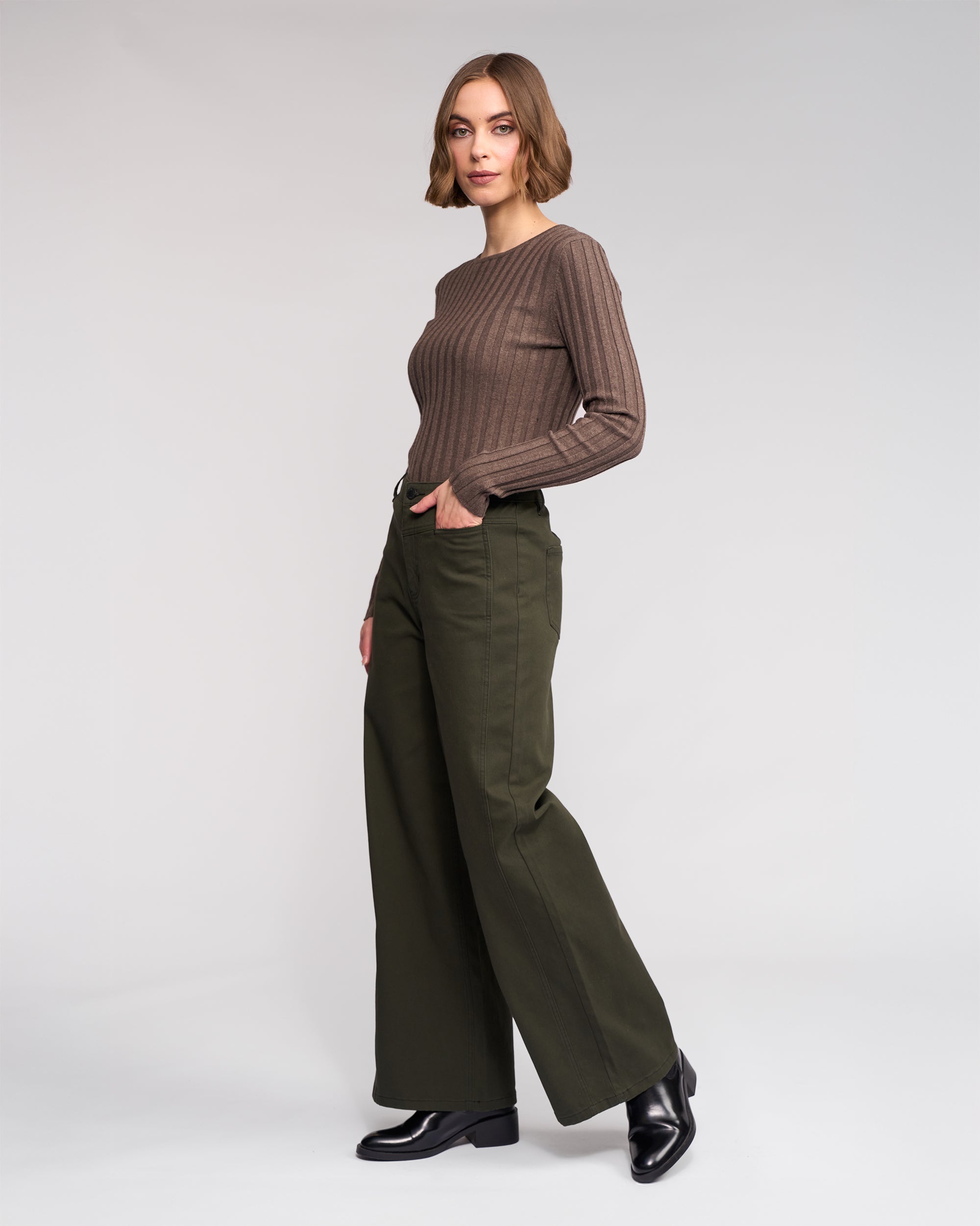 A woman stands against a plain backdrop, wearing Knewe Label's Hollie Jean - Hunter, paired with a long-sleeve brown ribbed sweater and black shoes. Her hands are in her pockets and she has a neutral expression.