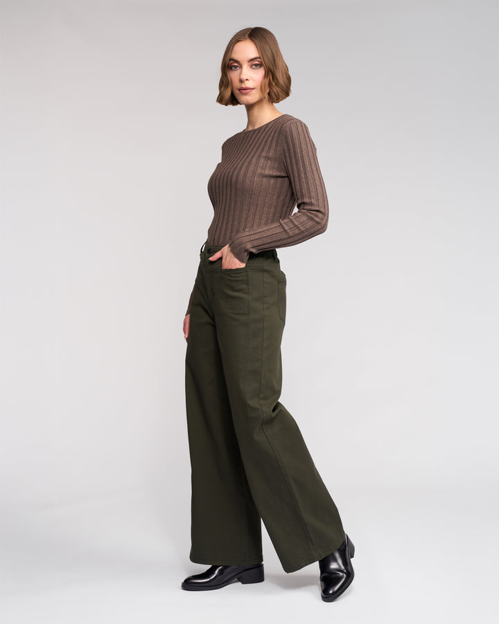 A woman stands against a plain backdrop, wearing Knewe Label's Hollie Jean - Hunter, paired with a long-sleeve brown ribbed sweater and black shoes. Her hands are in her pockets and she has a neutral expression.
