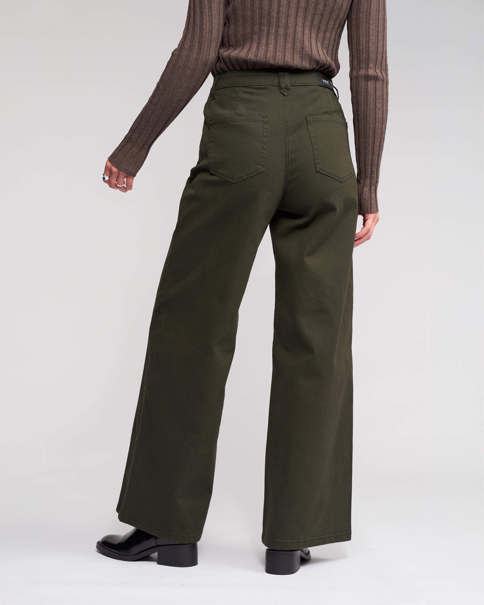 Wearing the Knewe Label Hollie Jean - Hunter, a person in a brown ribbed top and dark green wide-leg jeans with front welt pockets stands facing away against a light background, paired with black boots.