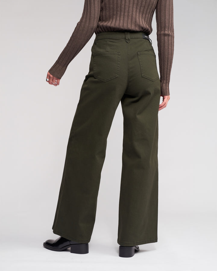 Wearing the Knewe Label Hollie Jean - Hunter, a person in a brown ribbed top and dark green wide-leg jeans with front welt pockets stands facing away against a light background, paired with black boots.
