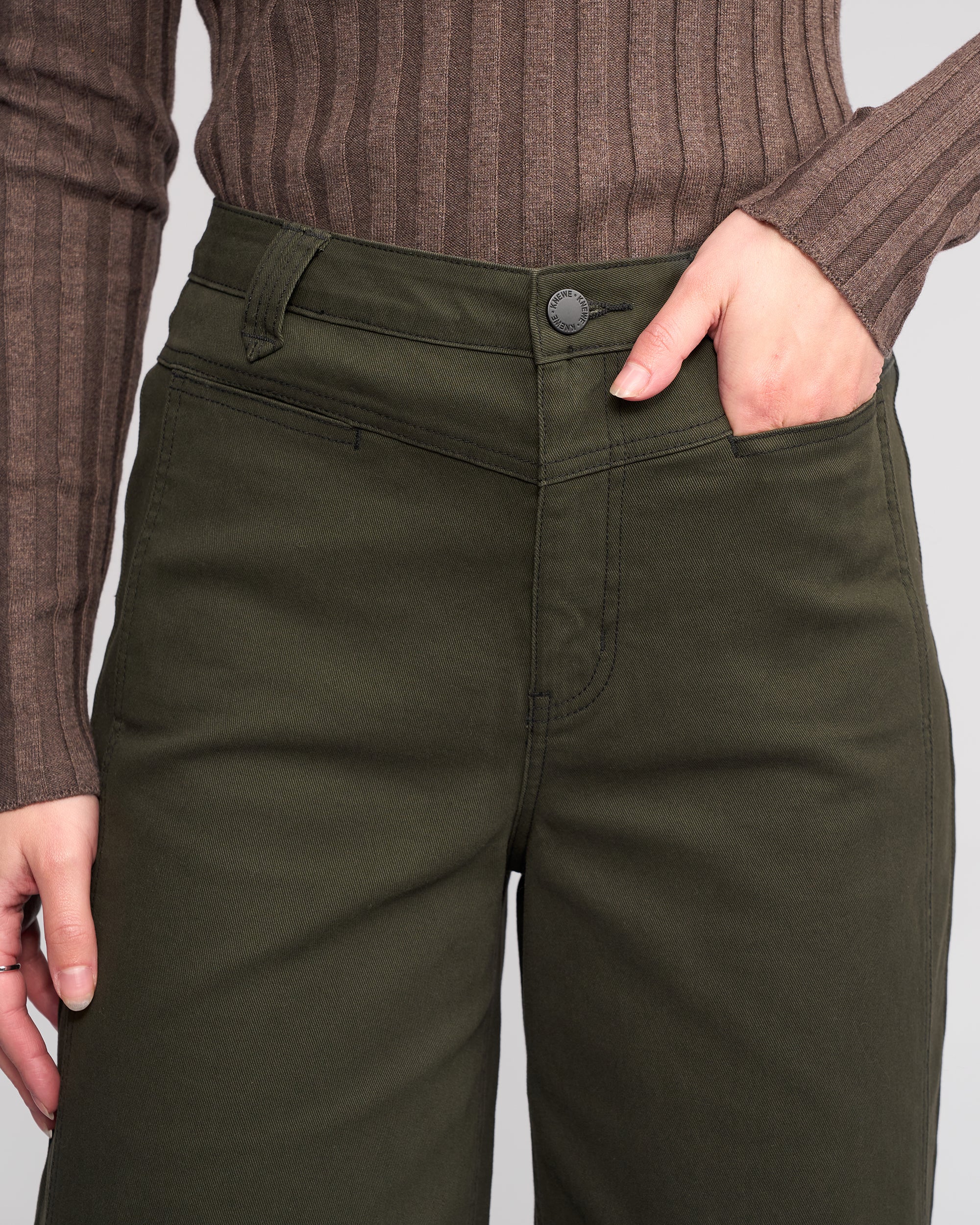 A person wears Knewe Label’s Hollie Jean - Hunter, dark green wide-leg jeans with front welt pockets and a brown ribbed sweater. Their right hand rests in a pant pocket. The image is cropped to show only the torso and upper legs.