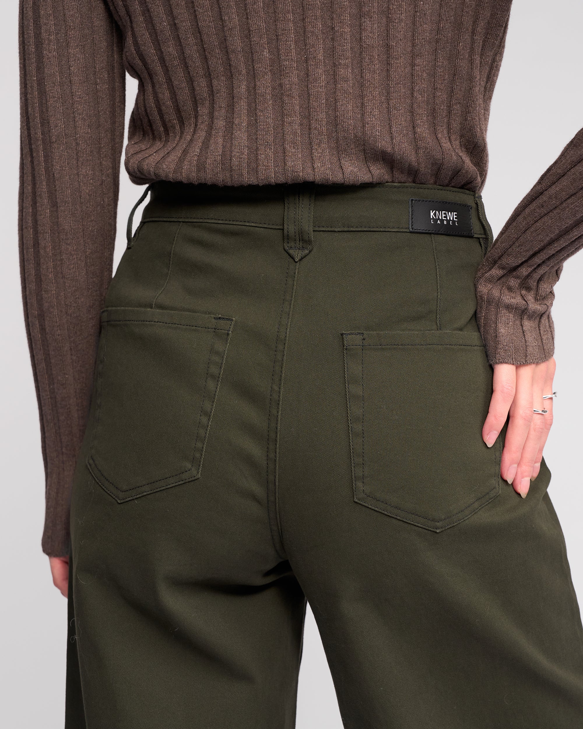 A person wearing the Hollie Jean - Hunter, a high-waisted dark green wide-leg jean by Knewe Label, and a brown ribbed sweater is shown from the back, one hand on hip with the black Knewe Label tag above the right pocket.