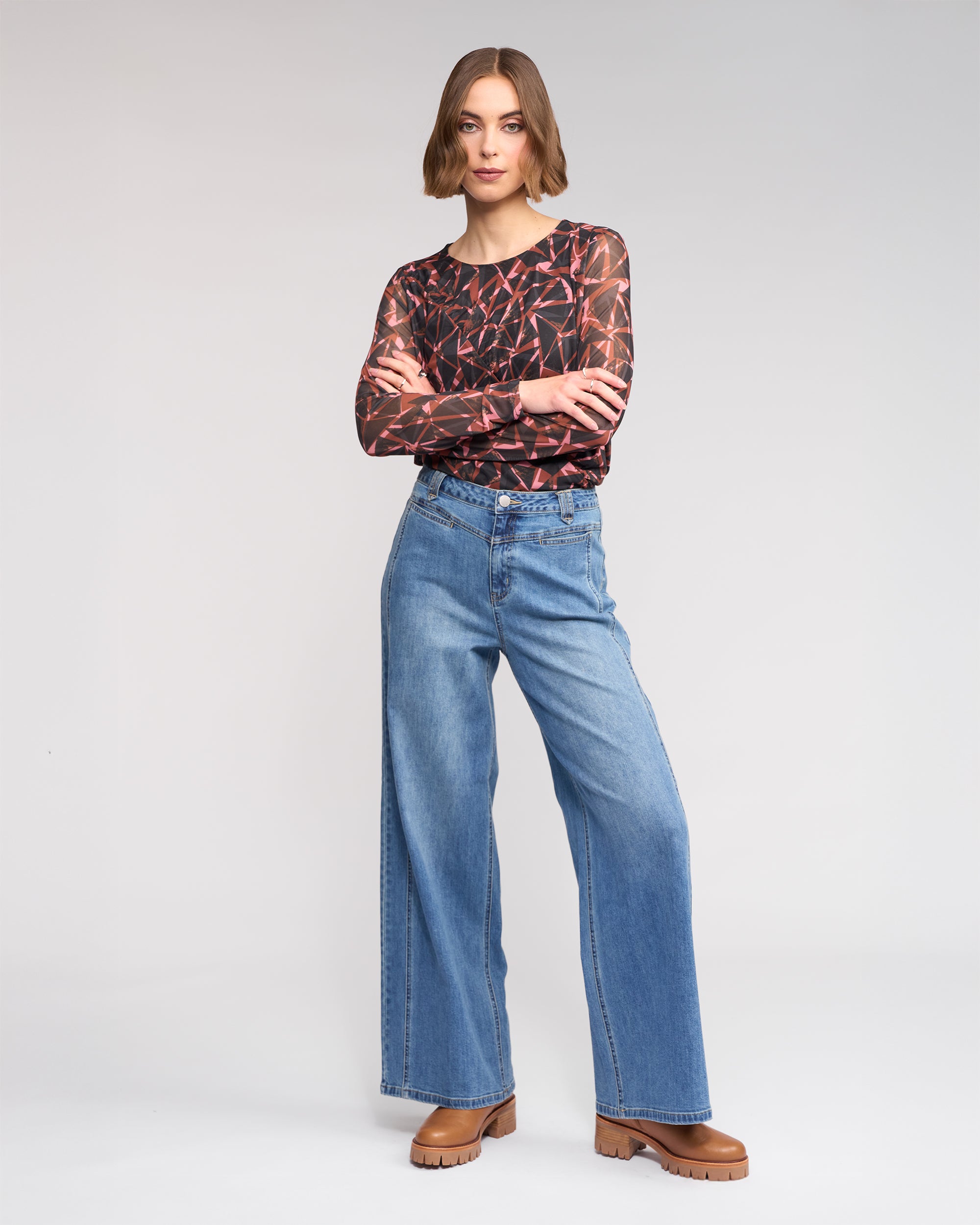 A woman with chin-length hair stands against a plain background wearing Knewe Label’s Hollie Jean - Vintage Denim, a long-sleeve abstract-patterned top, and brown platform shoes. She looks at the camera with arms crossed.