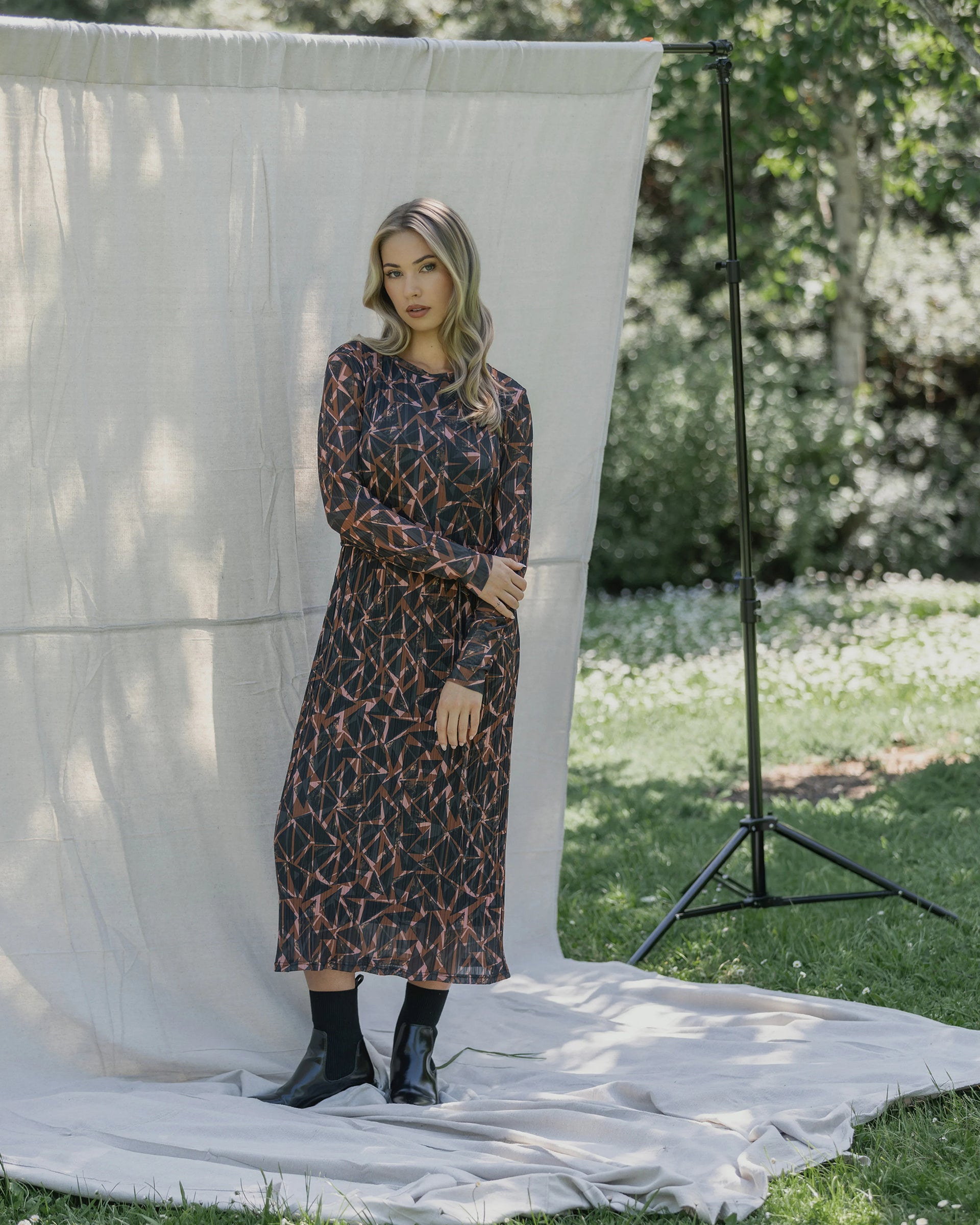 A woman stands outdoors on a white cloth before a neutral backdrop, wearing the Knewe Label Freddie Dress - Dusk with black boots, greenery and trees visible in the background.