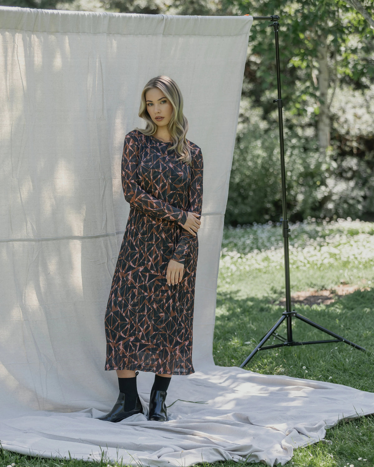A woman stands outdoors on a white cloth before a neutral backdrop, wearing the Knewe Label Freddie Dress - Dusk with black boots, greenery and trees visible in the background.