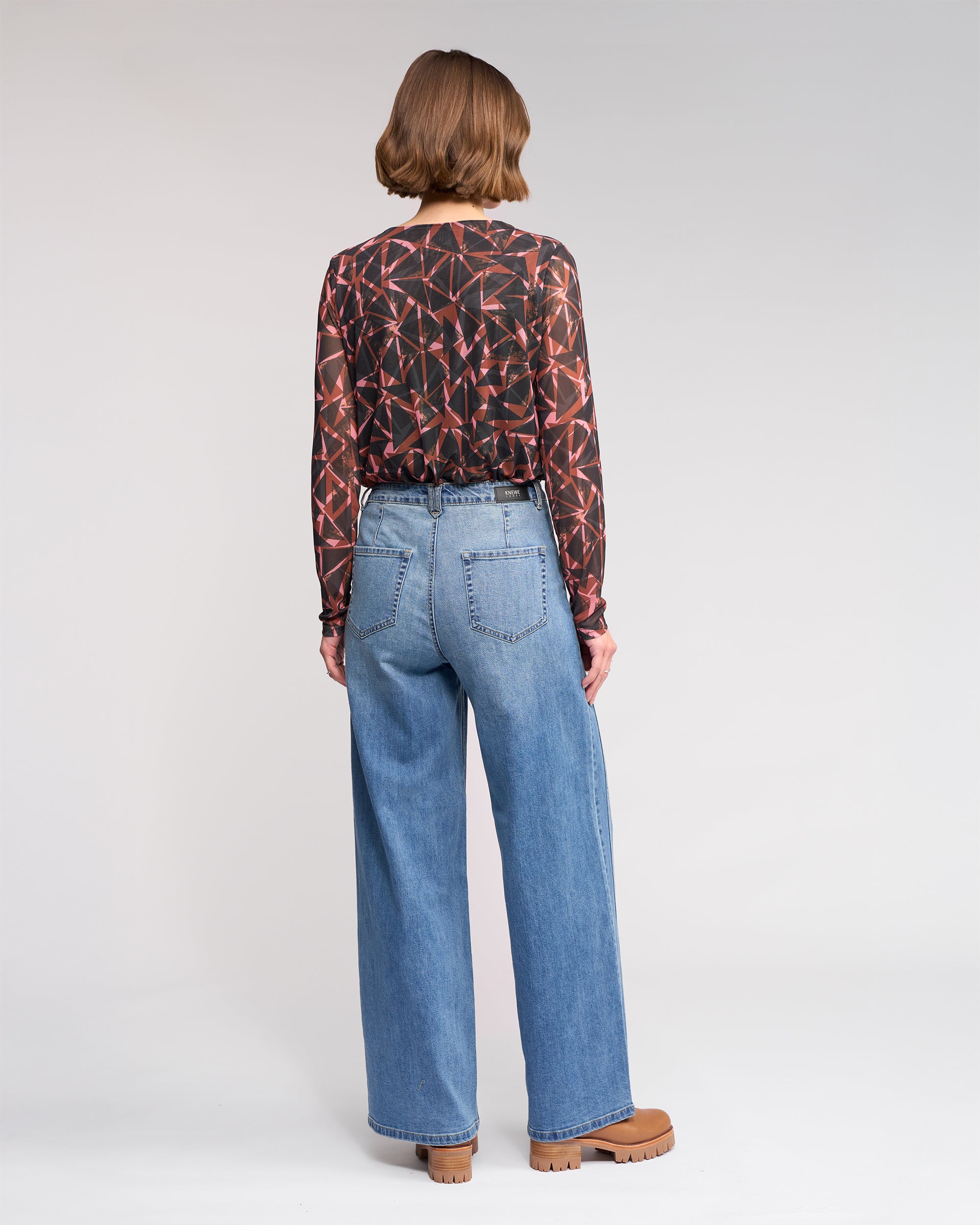 A person with chin-length brown hair stands facing away, wearing a geometric-patterned long-sleeve top, Knewe Label Hollie Jean - Vintage Denim in mid-blue, and brown platform shoes against a plain light background.