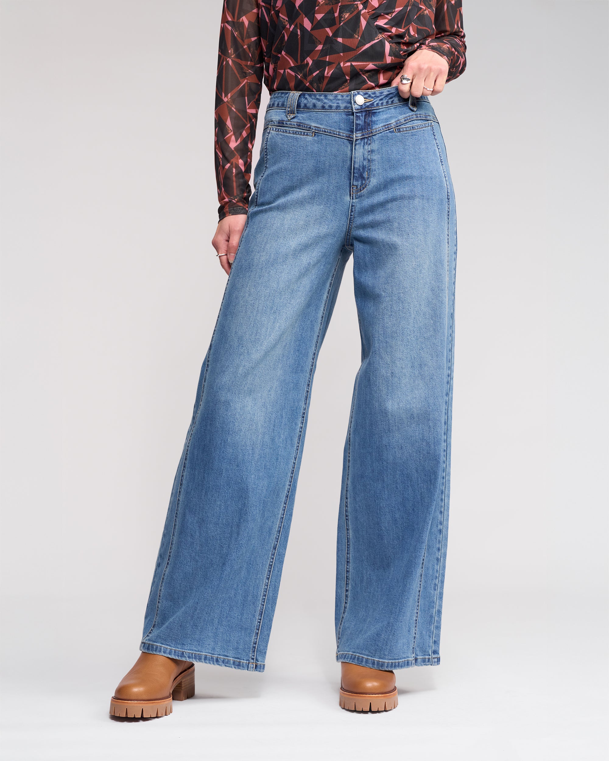 Model in Knewe Label’s Hollie Jean - Vintage Denim, styled with a geometric-pattern long-sleeve top and brown platform shoes, stands against a plain white background.