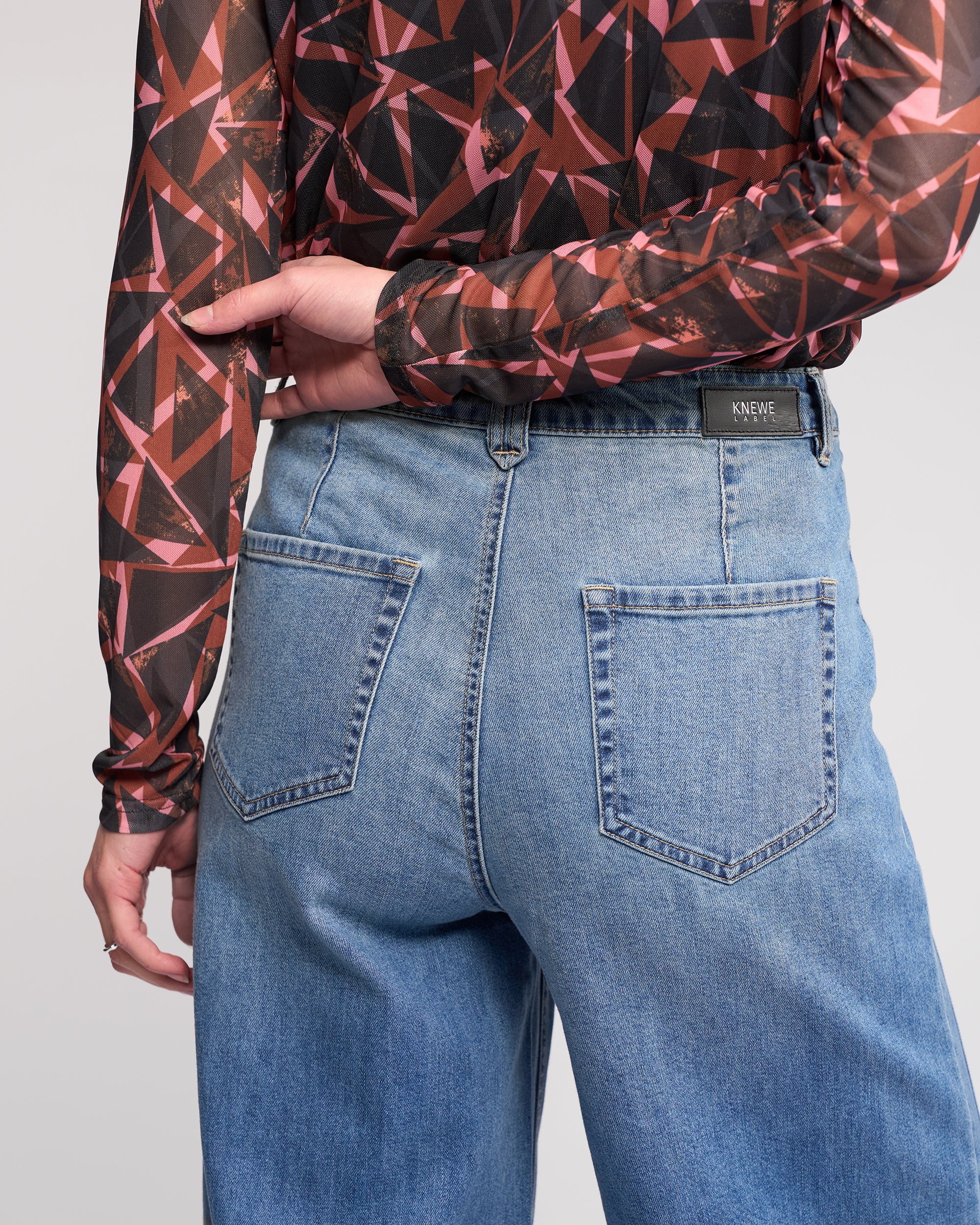 Wearing Knewe Label's Hollie Jean in vintage denim, a person stands with their back to the camera, showcasing high-waisted, mid-blue wide-leg jeans and a long-sleeved sheer geometric-patterned top.